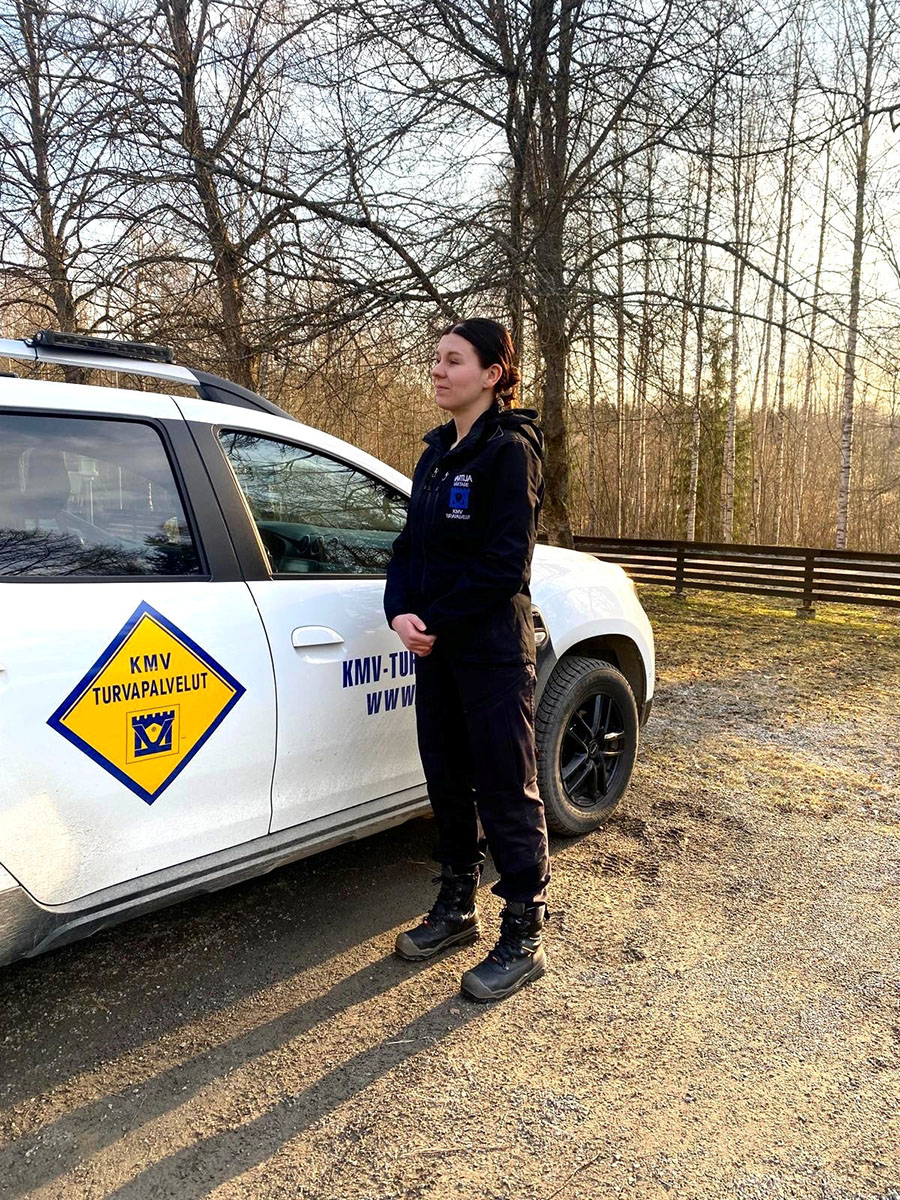 KMV Turvapalvelut_työntekijä Mustiin pukeutunut nainen seisoo valkoisen henkilötauton edessä, jonka kyljessä näkyy KMV-Turvapalveluiden logo.